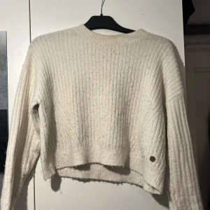 Vit ribbstickad tröja - Mysig vit ribbstickad tröja med rund halsringning och långa ärmar. Tröjan har en något croppad passform och en liten detalj i form av en knapp nertill. Perfekt för lager-på-lager under kyliga dagar.