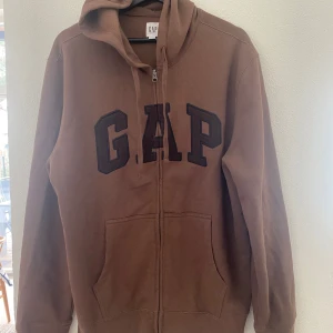 Brun hoodie med dragkedja från GAP - Säljer en brun hoodie från GAP i storlek L. Tröjan har dragkedja framtill, huva och stora fickor. GAP-loggan är broderad i svart på bröstet. Perfekt för en avslappnad stil.