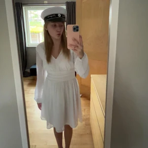 Vit v-ringad klänning med lång ärm - Säljer en vit klänning med v-ringning och långa ärmar. Klänningen har markerad midja och lätt utställd kjol som slutar ovanför knäna. Perfekt för student eller sommarens festligheter!