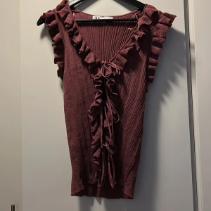 Vinröd ribbad topp med volang från Zara - Vinröd ribbad topp från Zara med djup v-ringning och volangdetaljer längs ärmhål och halsringning. Toppen har knyt framtill och är ärmlös. Säljer den då den är för stor för mig och har därmed inte använts💕