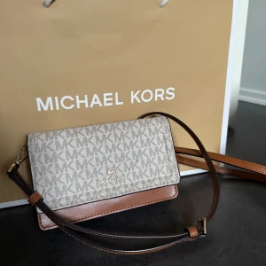 Beige och brun axelväska från Michael Kors - Snygg axelväska från Michael Kors i beige och brunt med MK-logga över hela framsidan. Väskan har justerbar axelrem, flera kortfack och dragkedjefack inuti. Perfekt för att bära det viktigaste med stil. Nypris:1995 kr. Använd ett fåtal gånger. Modell: Jet Set Charm Crossbody.