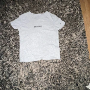 Ljusgrå t-shirt från Calvin Klein Jeans - Snygg ljusgrå t-shirt från Calvin Klein Jeans med svartvit logga på bröstet och ett litet 'CK'-broderi på ärmen. Klassisk rund halsringning och mjukt bomullsmaterial. Perfekt för en stilren och enkel look.