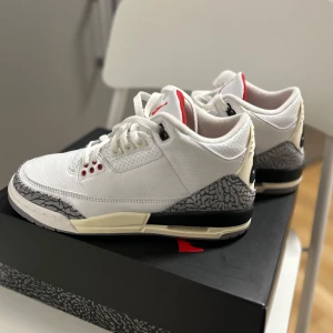 Nike Air Jordan 3 Retro White Cement - Säljer ett par Nike Air Jordan 3 Retro White Cement med vit ovandel i skinn, grått elefantmönster vid tå och häl, samt röda detaljer. Klassisk siluett med svart och beige sula och tydlig Nike Air-logga på hälen.