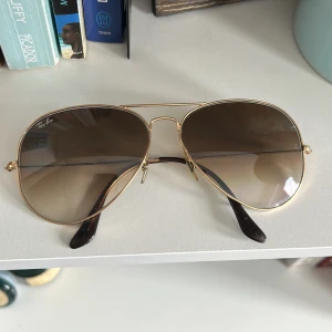 Ray-Ban aviator solglasögon med guldiga bågar - Snygga Ray-Ban aviator solglasögon med tunna guldiga metallbågar och bruna tonade glas. Klassisk pilotmodell som ger en cool och tidlös look. Skalmarna har mörka detaljer längst ut.