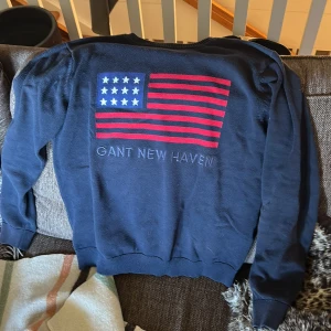 Mörkblå tröja med amerikansk flagga från Gant - Säljer en mörkblå tröja från Gant med stor amerikansk flagga och texten 'GANT NEW HAVEN' på ryggen. Tröjan har långa ärmar och ribbade muddar vid ärmslut och nederkant. Perfekt för dig som gillar klassisk stil.