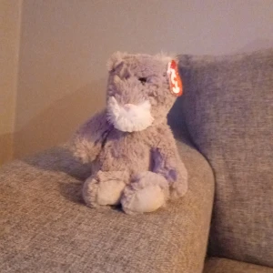 Grått gosedjur katt från Ty Beanie Babies - Säljer ett mjukt och fluffigt grått gosedjur i form av en katt från Ty Beanie Babies. Katten har vit nos och tassar samt ett rött hjärttag på örat. Perfekt för samlare eller som mysigt sällskap.