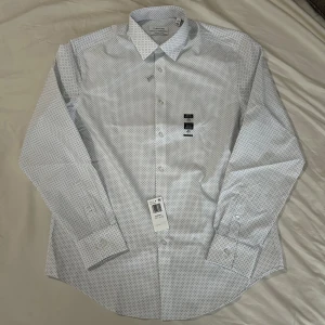 Calvin Klein skjorta ny - Säljer denna Calvin Klein skjortan som aldrig är använd och har lapparna kvar. Storlek L slimfit/34-35 i amerikansk storlek vilket motsvarar storlek XL/43. Nypris 599. Färgen är vit med små mörkblå och svarta detaljer. Priset på prislappen är borttagen på grund av att skjortan var en present. 