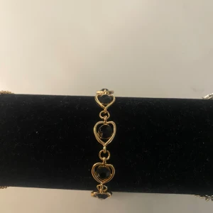 Guldfärgat armband med svarta hjärtan - Säljer ett elegant armband i guldfärg med flera öppna hjärtan och svarta stenar i hjärtform. Justerbar kedja för bra passform.priset går att diskutera💞