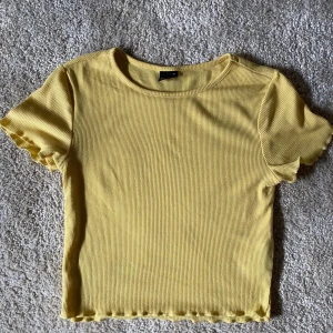 Gul ribbad croppad topp från Gina Tricot - Säljer en söt gul croppad topp från Gina Tricot. Toppen är ribbad med korta ärmar och har vågiga kanter vid ärmslut och nederkant. Perfekt för varma dagar och enkel att matcha med jeans eller kjol🌼💛