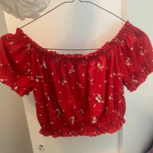 Röd blommig croppad topp - Superfin röd croppad topp med små blommor och resår vid axlar och midja. Toppen har korta puffärmar och en söt volangkant. Perfekt för varma dagar och ger en somrig känsla. Oanvänd