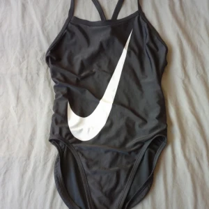 Nike baddräkt - Säljer en svart baddräkt från Nike med stor vit logga framtill. Baddräkten har smala axelband som korsas i ryggen och en öppen rygg för sportig look. Perfekt för simträning eller stranden. Står ingen storlek på, men skulle gissa på S. 