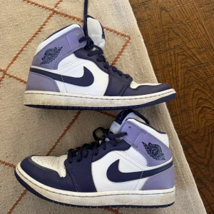 Air Jordan 1 Mid Blueberry - Säljer ett par Air Jordans i vitt och lila med hög siluett. Använda men i gott skick. Storlek 42.5. (cm 27). 1200kr. 