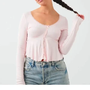 Ljusrosa croppad topp med spetskant - Söt ljusrosa croppad topp med lång ärm och dekorativ spetskant längs knäppningen framtill. Toppens material är mjukt och stretchigt, perfekt till jeans eller kjol.