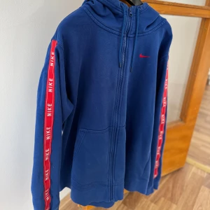 Blå hoodie från Nike med röda detaljer - Säljer en blå hoodie från Nike med dragkedja och huva. Tröjan har röda ränder med Nike-logga längs ärmarna och en liten röd Nike-swoosh på bröstet. Praktiska fickor framtill och justerbar huva med snören.