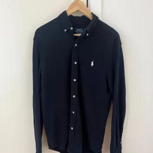 Marinblå skjorta från Polo Ralph Lauren - Klassisk marinblå skjorta från Polo Ralph Lauren med vit broderad logga på bröstet. Skjortan har långa ärmar, knappar framtill och en stilren krage. Perfekt för en clean och tidlös look.