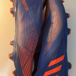Adidas Predator fotbollsskor blå och orange - Säljer ett par Adidas Predator fotbollsskor i blått med orange detaljer och mönster. Skorna har en strumpliknande passform utan snörning och är designade för maximalt grepp på planen. Perfekta för dig som vill sticka ut på fotbollsplanen.