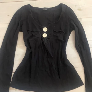 Svart långärmad topp med knappar från Gina Tricot - Säljer en svart långärmad topp från Gina Tricot med två stora dekorativa knappar framtill och lätt v-ringad hals. Toppen har figurnära passform och är perfekt för en stilren look.