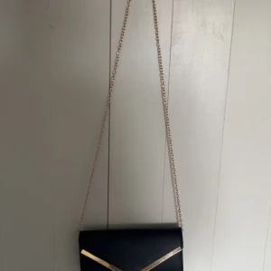 Svart kuvertväska med gulddetaljer och kedja - Snygg svart väska med en guldkedja som går att ta av och på. Fick i present men är inte riktigt min stil.