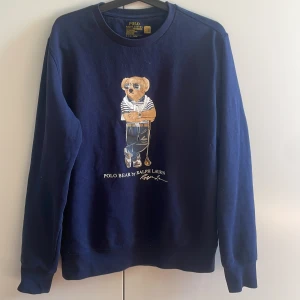 Marinblå sweatshirt med Polo Bear från Ralph Lauren - Säljer en marinblå sweatshirt från Ralph Lauren med den ikoniska Polo Bear-trycket framtill. Tröjan har rund halsringning och långa ärmar. Tillverkad i bomull och återvunnen polyester för en mjuk och bekväm känsla.