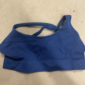 Blå ribbad sport-bh - Säljer en blå ribbad sport-bh med asymmetrisk design och smala axelband. Tillverkad i stretchigt material som består av 90% polyamid och 10% elastan. Perfekt för träning och ger bra stöd.