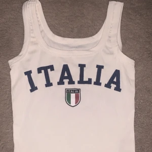 Vit ribbad linnetopp med ITALIA-tryck från SHEIN - Aldrig använt, säljer en vit ribbad linnetopp från SHEIN med blått ITALIA-tryck och Italiens flagga på bröstet. Toppen är ärmlös och tillverkad i 100% polyester. Perfekt för dig som gillar sportig stil eller vill visa ditt stöd för Italien.