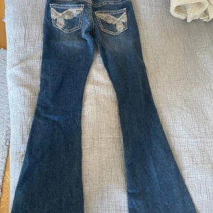 Gina Young 14+ jeans  - Snygga blå bootcut jeans med broderade detaljer på bakfickorna. Klassisk femficksmodell med låg midja och ljusa slitningar. Jeansen har en markerad söm och är perfekta för dig som gillar en lite retro vibe. Superlågmidjade och enbart använda 1 gång. Slutsålda på hemsida 