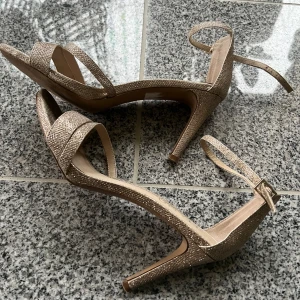 Glittriga guldiga klackskor med öppen tå - Säljer ett par glittriga guldiga klackskor från Only med öppen tå och smal rem runt vristen. Perfekta för att lyfta en outfit med sin eleganta design och höga klack. Skorna har en platt sula framtill och spänne vid ankeln. Storlek 41 men passar även 40!
