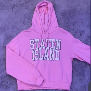 Rosa hoodie från H&M med tryck - Säljer en rosa hoodie från H&M med texten 'Staten Island New York City' i vitt tryck. Den har en klassisk huva med dragsko och långa ärmar. Perfekt för en avslappnad stil.