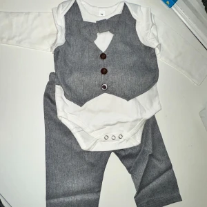 Grå babykostym med väst och fluga - Söt babykostym i grått med matchande väst och fluga. Setet inkluderar en vit långärmad body med knappar framtill och grå byxor med resår i midjan. Perfekt för speciella tillfällen! 6-12 månader Nyskick! 