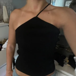 Svart oneshoulder topp - Snygg svart oneshoulder topp med en smal axelrem. Perfekt för en stilren och modern look. Passar bra till både jeans och kjol.