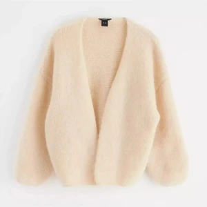 Beige stickad kofta - Mysig beige stickad kofta med långärmad design. Perfekt för kyligare dagar med en enkel och stilren look. Koftan har en unik knäppning framtill som ger en extra detalj.