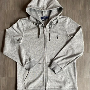 Grå hoodie från Polo Ralph Lauren - Säljer en stilren grå hoodie från Polo Ralph Lauren. Den har en dragkedja framtill och en broderad logotyp på bröstet. Tillverkad i mjuk bomull med ribbade muddar och justerbar huva. Perfekt för en avslappnad look.