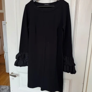 Svart klänning med volangärmar - Elegant svart klänning från Sand Copenhagen med långa ärmar och volangdetaljer vid ärmsluten. Perfekt för en stilren look. Klänningen har en klassisk och tidlös design. Storlek 40. Mycket fint skick!