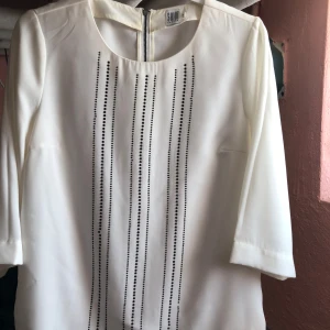 Vit blus med dragkedja från Soon - Elegant vit blus från Soon med dragkedja bak och dekorativa svarta pärlor framtill. Blusen har trekvartsärmar och en rund halsringning. Perfekt för en stilren look.