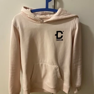 Ljusrosa hoodie från DirtCult - Säljer en ljusrosa hoodie från DirtCult med en stor ficka framtill och en tryckt logga på bröstet. Den har en klassisk huva och långa ärmar, perfekt för en avslappnad stil. Passar bra till både jeans och mjukisbyxor.