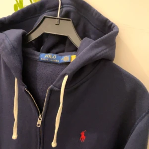 Mörkblå hoodie från Ralph Lauren - Säljer en snygg mörkblå hoodie från Ralph Lauren med dragkedja och vit snörning i huvan. Den har en röd broderad logga på bröstet och praktiska fickor framtill. Perfekt för en avslappnad stil. Kan passa S också! 