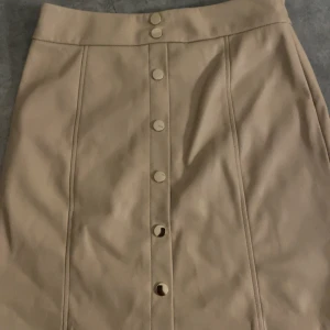 Beige tight skinn kjol  - Snygg beige kjol från New Yorker med knappar framtill. Kjolen har en stilren design och är perfekt för en chic look. Tillverkad i ett material som ger en skinnliknande känsla.