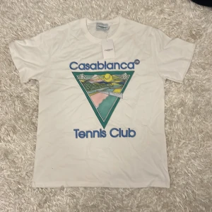 Vit t-shirt från Casablanca - Säljer en vit t-shirt från Casablanca med ett färgglatt tryck av ett landskap i en triangel på framsidan. Texten 'Casablanca Tennis Club' är tryckt i blått ovanför och under motivet. Perfekt för en avslappnad stil. Medela vid intresse 🤗