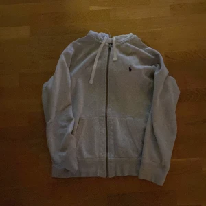 Grå hoodie från Ralph Lauren - Säljer en stilren grå hoodie från Ralph Lauren med dragkedja och justerbar huva. Den har en broderad logga på bröstet och praktiska fickor framtill. Perfekt för en avslappnad look. Litet hål i luvan men syns typ inte när man har den på