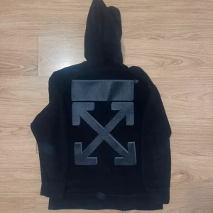 Svart hoodie från Off-White - Säljer en svart hoodie från Off-White med det ikoniska pilmönstret i grått på ryggen. Tröjan har en klassisk huva och en liten 'OFF' logga på framsidan. Perfekt för en stilren och modern look.
