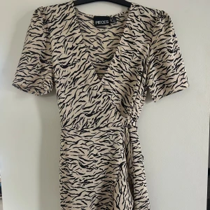 Beige omlottklänning från Pieces - Snygg beige omlottklänning från Pieces med svart zebramönster. Klänningen har korta ärmar och ett knytband i midjan för en smickrande passform. Använd ett flertal gånger men är fortfarande i ett fint skick, finns en liten svart märke på ryggen (se sista bild). Som inte syns på långt håll. Hör av dig om du vill ha fler bilder eller har frågor:)