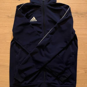 Mörkblå träningsjacka från Adidas - Säljer en mörkblå träningsjacka från Adidas med dragkedja och vita detaljer. Jackan har långa ärmar och en klassisk sportig look. Perfekt för träning eller en avslappnad stil. Adidas-loggan syns tydligt på bröstet.