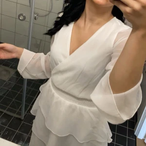 Vit omlottklänning - Säljer en elegant vit omlottklänning med v-ringning och långa ärmar. Klänningen har volangdetaljer och ett elastiskt band i midjan för en smickrande passform. Perfekt för sommarens festligheter!