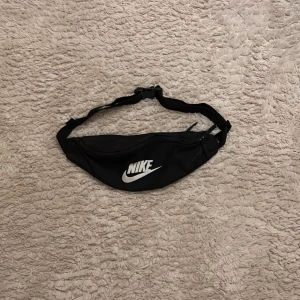 Svart midjeväska från Nike - Snygg och praktisk svart midjeväska från Nike med justerbart band och dragkedja. Perfekt för att hålla dina saker säkra och nära till hands. Den har ett stort fack med meshficka inuti för extra förvaring.