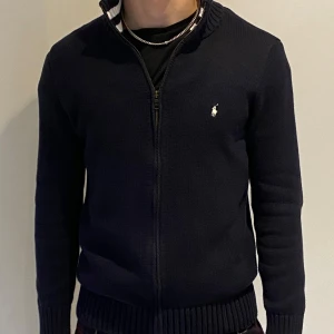 Svart stickad tröja från Ralph Lauren - Säljer en stilren svart stickad tröja från Ralph Lauren med dragkedja och ribbade muddar. Tröjan har en liten broderad logotyp på bröstet och långa ärmar. Perfekt för en klassisk look. Nästan helt ny använd fåtal gånger,Storlek S