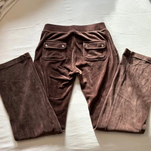 Bruna mjukisbyxor från Juicy Couture - Snygga bruna mjukisbyxor från Juicy Couture med snörning i midjan och praktiska fickor med knappar bak. Perfekta för en avslappnad stil med en mjuk och bekväm känsla. Jätte bra skick på sametstyget då byxorna endast varit använde 2-3 gånger och tvättade kanske en gång