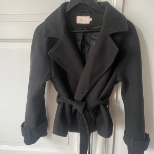 Svart kappa från Nelly - Snygg svart kappa från Nelly med knytskärp i midjan och breda slag. Jackan har långa ärmar med knappar vid ärmsluten och en stilren design som passar perfekt för kyligare dagar.
