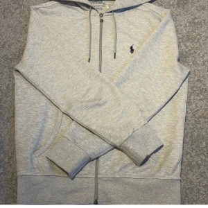 Grå hoodie från Polo Ralph Lauren - Säljer en stilren grå hoodie från Polo Ralph Lauren med dragkedja och huva. Tröjan har långa ärmar och en liten broderad logga på bröstet. Perfekt för en avslappnad look. Den är bara använd 3 gånger, ny pris kostar runt 2000kr!  