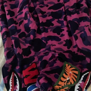 Lila camo hoodie från A Bathing Ape - Säljer en cool lila camo hoodie från A Bathing Ape med dragkedja och hajmotiv på huvan. Perfekt för dig som vill sticka ut med en unik stil. Hoodien har en iögonfallande design med mörka och ljusa lila nyanser.