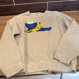 Grå sweatshirt  - Säljer en grå sweatshirt från Uniqlo i samarbete med KAWS. Tröjan har ett unikt tryck med en blå figur och en gul banan på framsidan. Tillverkad i en mjuk bomulls- och polyesterblandning. Perfekt för dig som gillar konstnärliga och lekfulla motiv! Helt ny